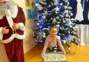 Uśmiechnięta dziewczynka trzyma w ręku prezent, stoi obok figury Mikołaja.